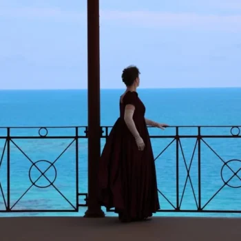 A woman in a red dress stands on a balcony, gazing out at the vast ocean below.
