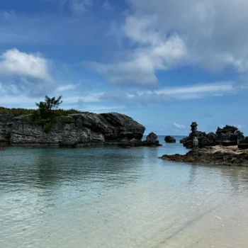 A serene sandy beach featuring scattered rocks, with calm water visible in the distance under a clear sky.